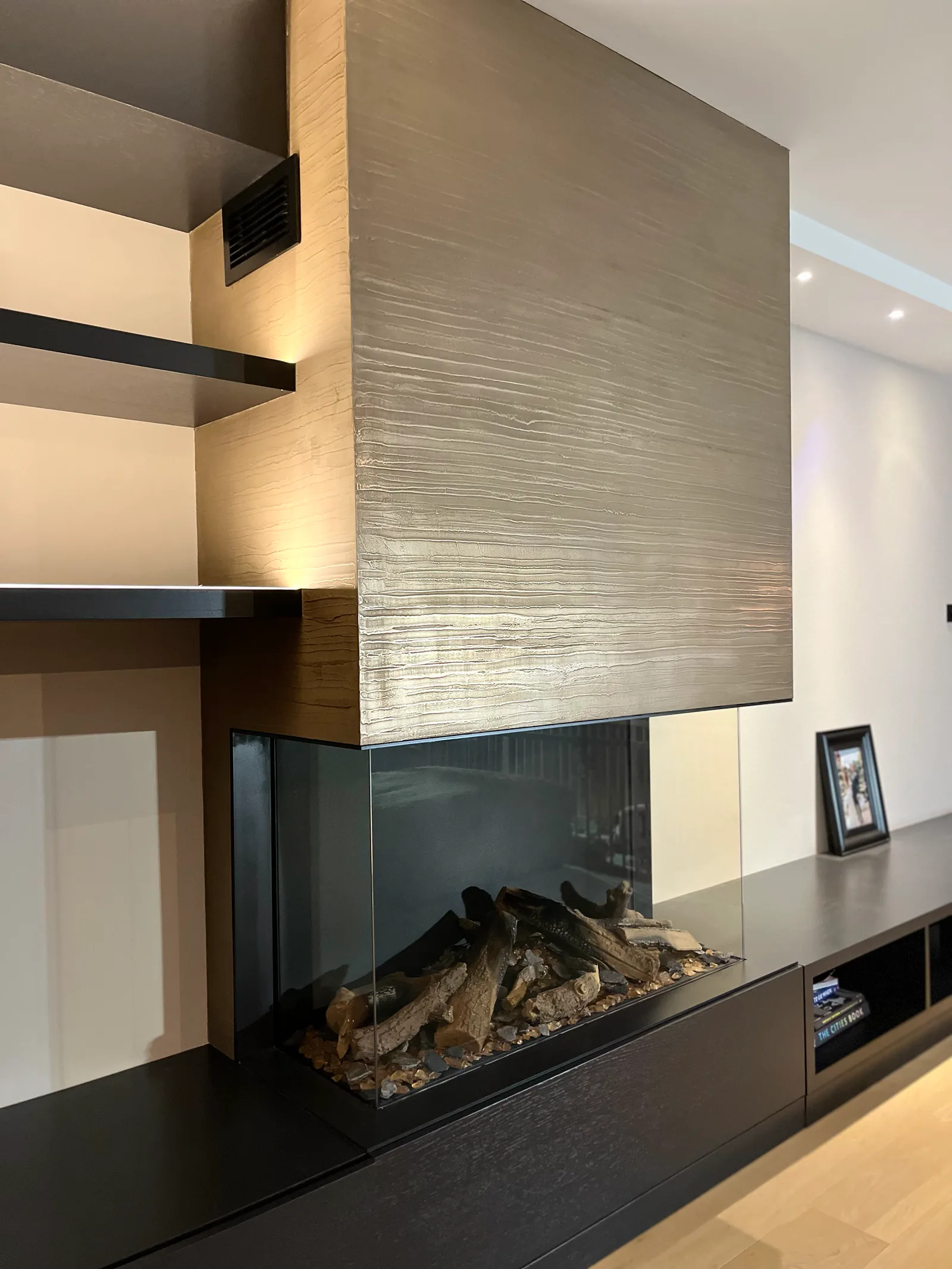 Een haardombouw met horizontaal reliëf, een glazen hoekhaard en een donker meubel met open planken.