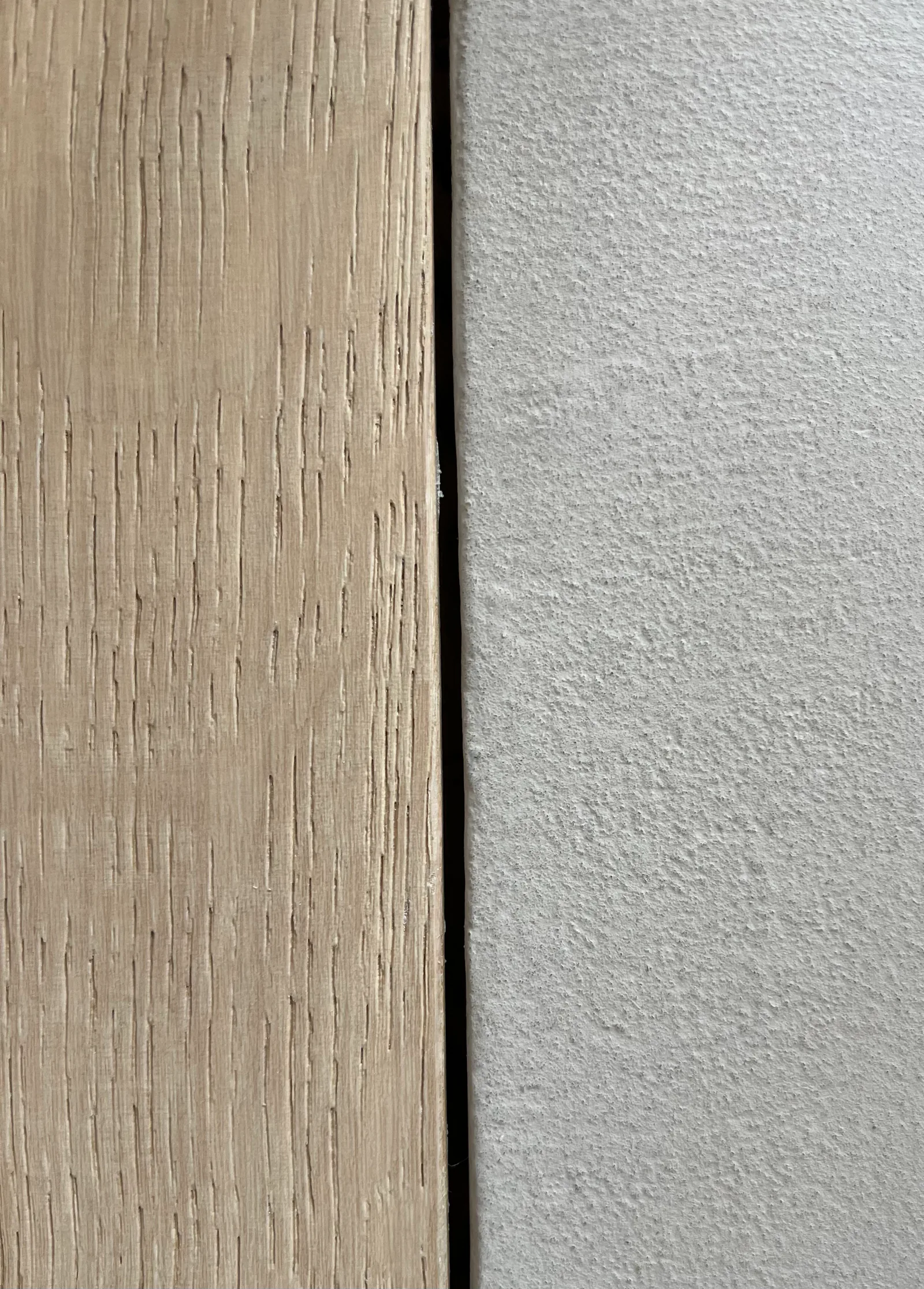 Detail van een houten paneel naast een lichtgrijs getextureerd oppervlak, gescheiden door een smalle donkere voeg.