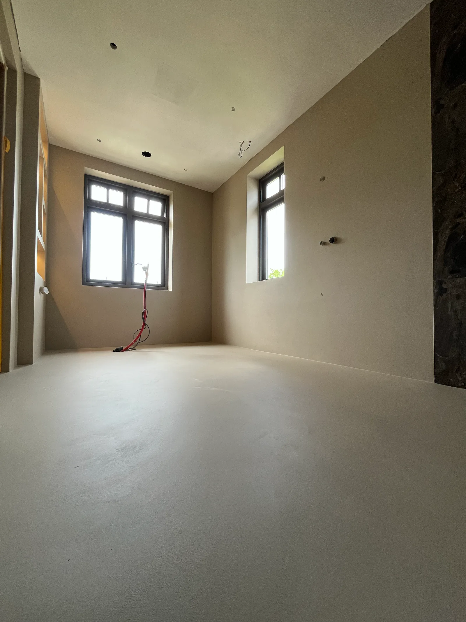 Een lege ruimte met een lichte doorlopende vloer, donkere raamkozijnen en onafgewerkte aansluitpunten.